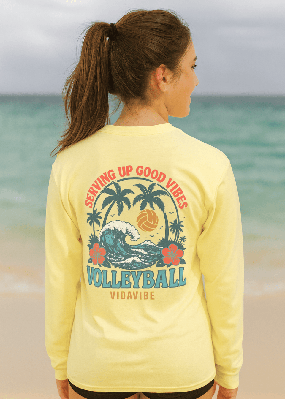Woman wearing a yellow long-sleeve shirt with volleyball design and text on a beach.